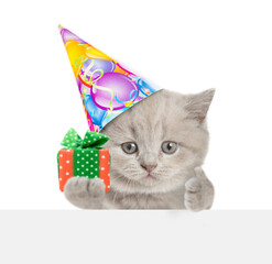 Poster - Funny tiny kitten wearing party cap holding gift box above blank white banner and showing thumbs up gesture. isolated on white background