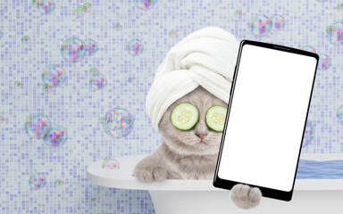 Leinwandbilder - Kitten with towel on it head, with pieces of cucumber on it eyes and with cream on it face takes the bath at home and shows big smartphone with white blank screen in it paw.  Empty space for text