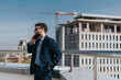 © qunica.com - Confident businessman in a suit standing on a balcony, making a phone call. The background features a construction site, highlighting themes of business communication and urban development.