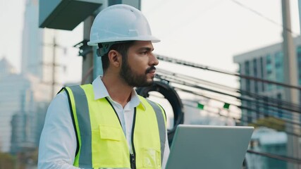 Portrait of electrician working on laptop near power transmission lines. Bearded engineer checks electrical network settings at construction site. Modern building technology