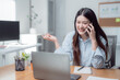 © MINAE - Young Asian businesswoman talking on phone and working on laptop, smiling and gesturing while sitting at desk in modern office, enjoying her successful freelance career