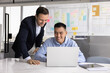 © fizkes - Happy Asian business professional man showing work result to boss standing nearby, sitting at laptop, typing, smiling. Multiethnic colleagues analyzing online product on computer