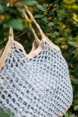 Handmade gray bag crocheted in the form of a net against the background of a summer bush. Author's handmade accessories. Crochet as a way to relax and earn money.