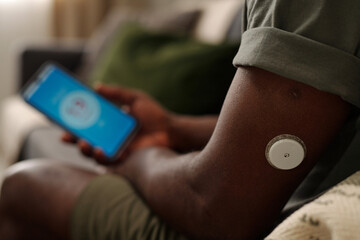 Close-up of person using smartphone for blood glucose monitoring showing patch on arm with focus on gadget in hand and background blurred