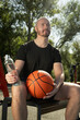 © svetlana_cherruty - Basketball player or sports man in black t-shirt sitting and resting with water bottle and basketball orange ball after game. Basketball coach. Outdoors. Water balance for heat season.