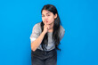© Saddha Visual - Asian young woman with long black hair, wearing a gray shirt, playfully making a shushing gesture with her finger on lips, smiling mysteriously against a blue background.