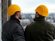 © NawapDesign - Two Construction Workers in Yellow Hard Hats Discussing Project Progress Overlooking City Skyline