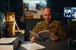 © DragonImages - Portrait of an army officer intently reviewing confidential documents in a dimly lit office setting. Background includes various military-themed posters and a computer setup