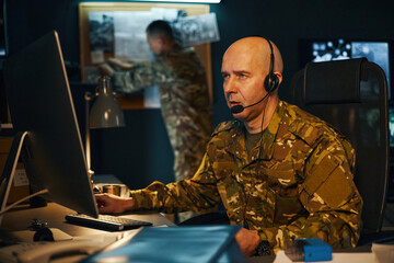 Man in military uniform using computer with headset on, focused on task with another person in the background engaging with equipment