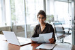 © Stock 4 You - Smiling portrait at camera business woman accountant holding documents, working at laptop pc computer doing online tax invoice, financial report. Latina businesswoman manager with paperwork in office