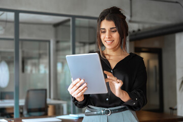 Latina Hispanic young adult professional business woman looking at pc device computer screen. Indian female businesswoman finance specialist holding digital tablet using fintech tab standing in office