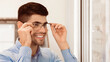 © Prostock-studio - A young man smiles as he adjusts a pair of stylish glasses while standing in a well-lit eyewear store. Natural light enhances the cheerful atmosphere of his experience.