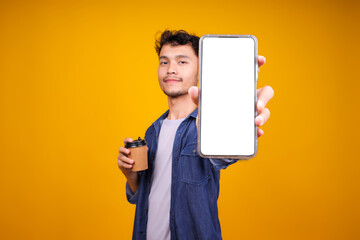 Asian Guy Holding Coffee Cup And Showing Smartphone With White Blank Screen For Mockup Over Yellow Background