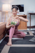 © Tj - Happy asian fitness girl in sportswear sitting on yoga mat and eating a fresh vegetable salad after workout at home, healthy food and lifestyle concept