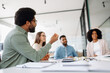 © Vadim Pastuh - Confident businessman in casual attire sitting at a desk with a smile. Modern office setting with a productive team in the background discussing projects and ideas.