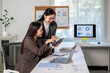 © Wasana - Two women are sitting at a desk with a laptop and tablet in front of them