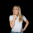 © Iryna - Blonde woman keeps hand on chin looks pensively above dressed in casual white t-shirt poses against black background blank copy space for your advertising content thinks about future