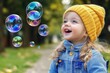 © Michael - Happy toddler girl wearing yellow beanie and denim jacket watching soap bubbles floating in the air in a park