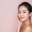 © julimur - Young Asian woman with flawless skin gently touching her cheek, smiling softly against a pastel pink background. Natural beauty, skincare concept.