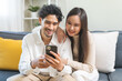 © KMPZZZ - Happy couple love at home, two asian young spending time, bonding to each other romantic on sofa in living room while man embrace woman using smartphone, mobile phone to selfie take a photo together.