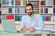 © Valerii Honcharuk - Young male teacher working in library sitting at desk with laptop books, looking at camera