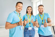 © Studio Romantic - Group of young cheerful volunteers in blue t-shirts standing together, showing thumbs up, looking at camera. Positive men and woman working in charity center, providing humanitarian help and charity.