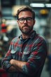 © Aleksander - A industrial designer man with glasses and a beard stands confidently with arms crossed, wearing a plaid shirt. The background is a blurred workshop setting, suggesting a skilled tradesman.