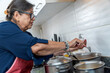 © FABIAN PONCE GARCIA - Senior woman adding ingredients while cooking soup in kitchen