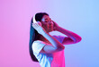 © Prostock-studio - A young Asian woman with long black hair is deeply immersed in music while wearing headphones. Soft pink and blue lights create a vibrant atmosphere as she closes her eyes in contentment.