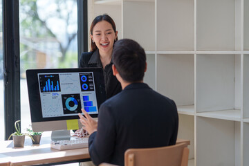 Two asian businesspeople are discussing business performance using charts and data displayed on a computer screen, working together in the office