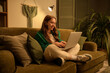 © Pixel-Shot - Young woman using laptop on sofa at home in evening