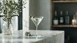 © Elena - Martini glass on a marble countertop in a kitchen. the glass is filled with a clear liquid and has two olives on the rim. on the left side of the countertop, there is a small vase with green leaves.