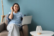 © Pixel-Shot - Young woman using laptop in grey armchair near blue wall at home