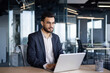 © Liubomir - Serious thinking businessman working with laptop inside office. Man in business suit sitting at desk typing on keyboard, working late into the night.