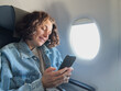 © Юлия Завалишина - Passenger woman is flying in plane. Girl using phone sitting by airplane window. Traveling female with smartphone inside plane enjoying flight. Woman using wifi connection on board. Traveling girl
