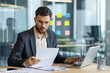 © Liubomir - Serious thinking businessman behind paperwork, man checking financial and investment reports. Financier in business clothes holding papers and documents in hands, focused inside office.