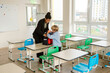 © Drobot Dean - A Black female teacher in a black blazer helps a young White boy in a blue shirt pack his backpack in a bright classroom. Green and blue chairs are arranged around desks.