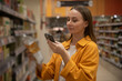 © Grustock - In a grocery store, a woman scans a coffee package with her phone while browsing the aisles. She appears focused on her shopping task, selecting products carefully.