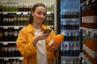 © Grustock - In a grocery store, a woman dressed in a yellow jacket scans a bottle of orange juice using her mobile device, surrounded by various beverages on shelves.