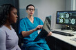 © pressmaster - Doctor wearing glasses holding MRI scan and explaining results to patient in medical office. Multiple screens showing medical data on desk