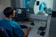 © pressmaster - Medical professional in blue scrubs monitoring brain scans on computer screen while patient lies in MRI machine. Clear view of medical equipment and technology