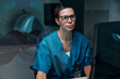 © pressmaster - Portrait of Caucasian medical professional wearing glasses and blue scrubs working with high-tech medical equipment at healthcare facility