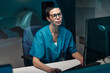 © pressmaster - Portrait of female doctor with glasses wearing scrubs, working on computer, examining medical data in office. Blue medical equipment seen in background