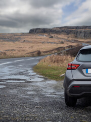 Wall Mural - A car is parked on a wet road with a view of a mountain in the background. The car is a grey. Travel to nature by car concept. Burren Ireland. Adventure with rented vehicle.