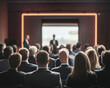 © Inspire Shots Hub - Audience viewed from behind, seated in a dark room before a screen and speaker.  Illustrates a business conference, presentation, or seminar setting.  Showcase of event space.