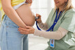 © New Africa - Pregnancy checkup. Doctor with stethoscope listening baby's heartbeat in patient's tummy indoors, closeup