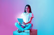 © Prostock-studio - Asian woman sits cross-legged on a cube in a creative space, expressing surprise while using a tablet. The vibrant blue and pink lighting adds a dynamic atmosphere to the moment.