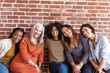 © Rawpixel.com - Group of five diverse women sitting together, smiling in front of a brick wall. Diverse ethnicities and different ages, casual attire, friendly atmosphere, cheerful expressions.