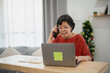 © WMSTUDIO - Senior Asian woman using laptop while talking on smartphone at home, enjoying a peaceful moment in the modern living space with holiday decorations