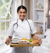 © wavebreak3 - Serving breakfast tray with coffee and fruits, healthcare professional smiling at home